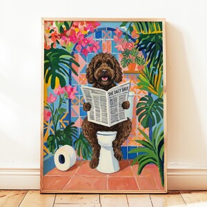 Op de afbeelding: Een ingelijste kunstprint toont een bruine hond op een toilet, lezend in de krant "The Daily Bark". De achtergrond bevat weelderig groen, roze bloemen en een blauwe tegelmuur. Een rol toiletpapier ligt op de vloer.