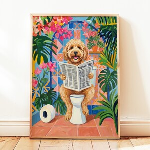Könnte beinhalten: Ein gerahmter Druck zeigt einen Golden Doodle Hund auf einer Toilette, der die Zeitung "The Daily Bark" liest. Der Hintergrund zeigt eine farbenfrohe Badezimmerszene mit tropischen Pflanzen und floralen Akzenten. Eine Rolle Toilettenpapier ist sichtbar.