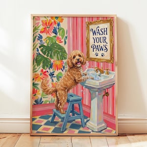 Op de afbeelding: Een ingelijste kunstprint met een goudbruine hond die op een blauw opstapje staat en zijn poten wast in een witte wastafel op voet. De achtergrond toont een bloemenbehang en een goudkleurig bord met de tekst WASH YOUR PAWS.