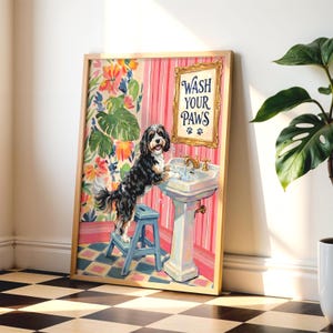 Pode incluir: Uma impressão de arte emoldurada apresentando um cão preto e branco em pé num banco azul para lavar as patas num lavatório branco. O fundo mostra papel de parede às riscas cor-de-rosa, padrões florais e uma placa dourada com o texto escrito.
