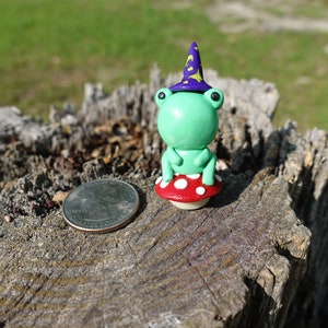 May include: A green frog wearing a purple and yellow wizard hat sits on a red and white mushroom. The frog and mushroom are made of polymer clay and are about the size of a quarter.