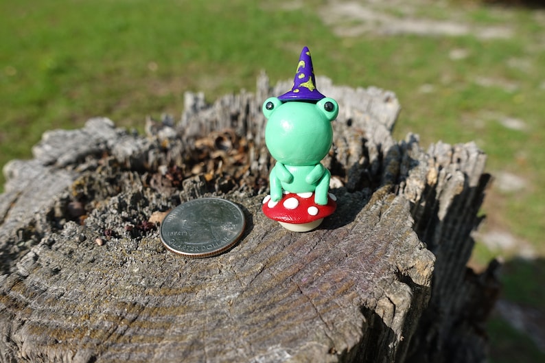 May include: A green frog wearing a purple and yellow wizard hat sits on a red and white mushroom. The frog and mushroom are made of polymer clay and are about the size of a quarter.
