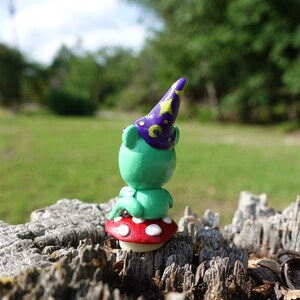 May include: A small green figurine wearing a purple hat with gold stars sits on a red and white mushroom. The figurine is sitting on a tree stump.