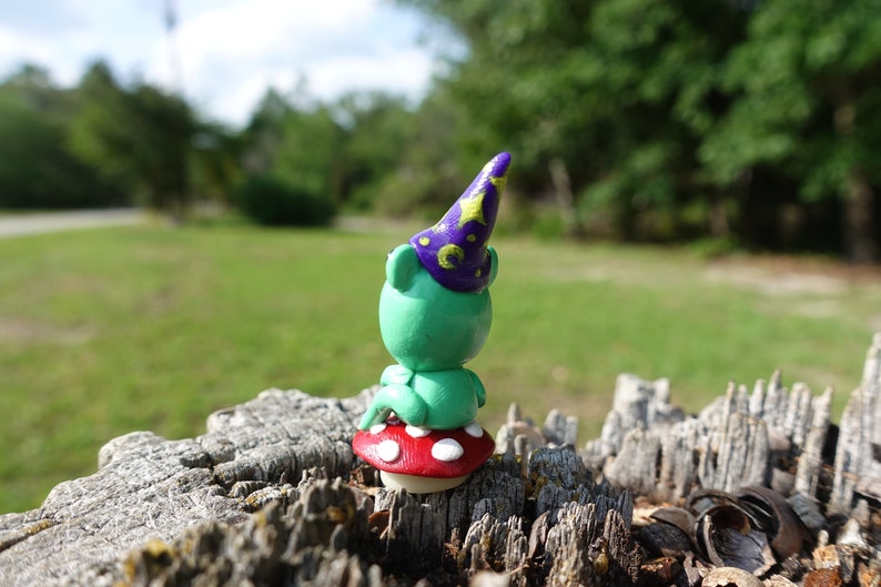 May include: A small green figurine wearing a purple hat with gold stars sits on a red and white mushroom. The figurine is sitting on a tree stump.