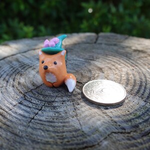 May include: A small orange fox figurine wearing a teal and pink hat with a white tail. The figurine is sitting on a wooden surface.