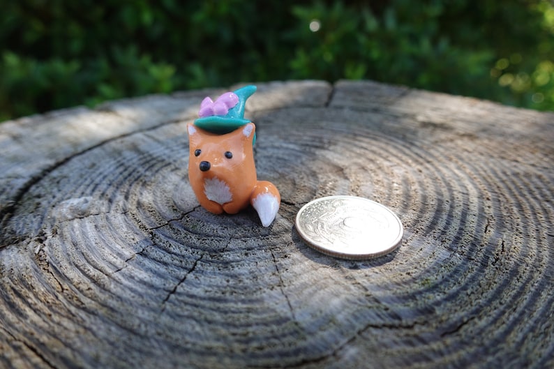 May include: A small orange fox figurine wearing a teal and pink hat with a white tail. The figurine is sitting on a wooden surface.