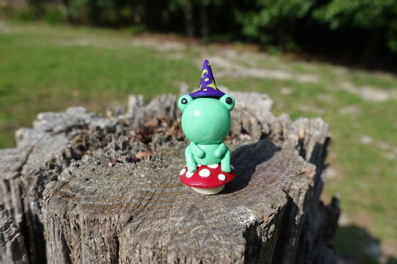 May include: A green frog wearing a purple and yellow wizard hat sits on a red and white mushroom. The frog and mushroom are made of clay and are sitting on a tree stump.