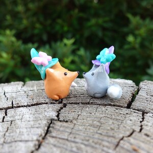 May include: Two small clay figurines of foxes wearing flower crowns. The fox on the left is orange and the fox on the right is gray. They are both looking at each other.