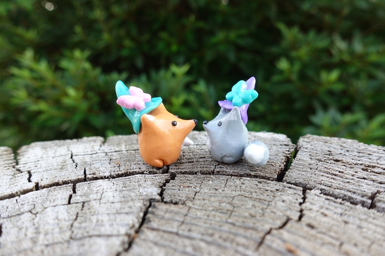 May include: Two small clay figurines of foxes wearing flower crowns. The fox on the left is orange and the fox on the right is gray. They are both looking at each other.