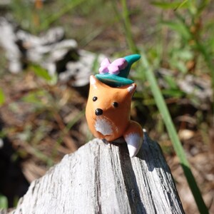 May include: A small orange fox figurine wearing a teal blue witch hat with a pink bow. The figurine is sitting on a piece of weathered wood.