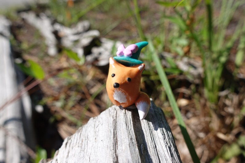 May include: A small orange fox figurine wearing a teal blue witch hat with a pink bow. The figurine is sitting on a piece of weathered wood.