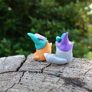 May include: Two small clay figurines of foxes wearing hats. The fox on the left is orange and wearing a teal hat with a pink flower. The fox on the right is gray and wearing a purple hat with a teal flower.