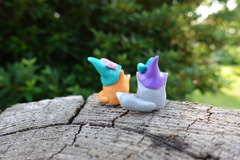 May include: Two small clay figurines of foxes wearing hats. The fox on the left is orange and wearing a teal hat with a pink flower. The fox on the right is gray and wearing a purple hat with a teal flower.