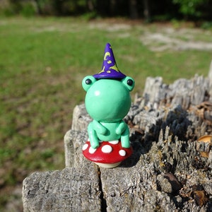 May include: A green frog wearing a purple and gold wizard hat sits on a red and white mushroom. The frog is made of polymer clay and has black eyes.
