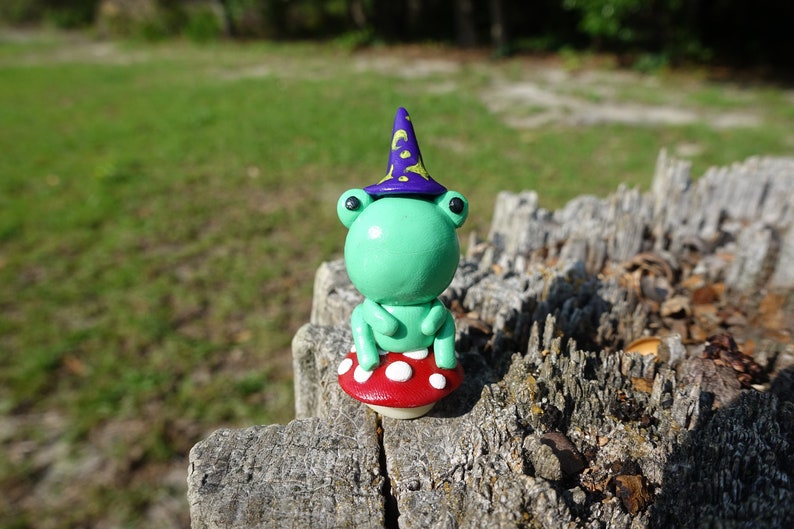 May include: A green frog wearing a purple and gold wizard hat sits on a red and white mushroom. The frog is made of polymer clay and has black eyes.