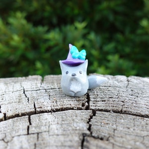 May include: A small gray wolf figurine wearing a purple witch hat with a teal butterfly on top. The figurine is sitting on a weathered wooden surface.