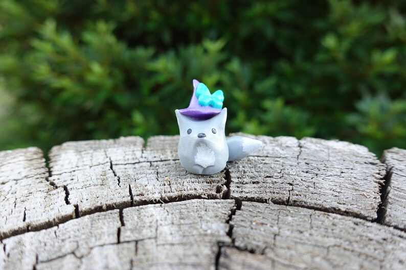 May include: A small gray wolf figurine wearing a purple witch hat with a teal butterfly on top. The figurine is sitting on a weathered wooden surface.