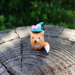 May include: A small orange fox figurine wearing a teal blue hat with a pink bow. The figurine is sitting on a wooden surface.