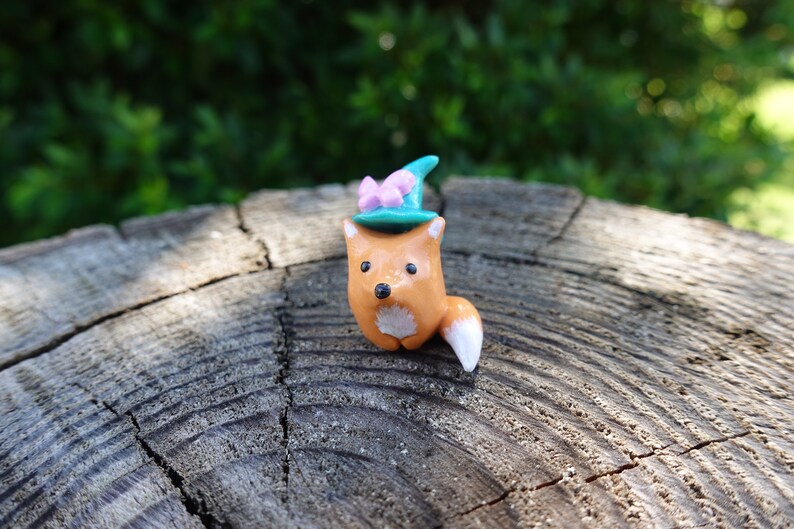 May include: A small orange fox figurine wearing a teal blue hat with a pink bow. The figurine is sitting on a wooden surface.