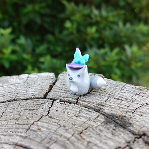 May include: A small gray clay figurine of a fox wearing a purple and blue witch hat with a blue butterfly on top. The figurine is sitting on a gray wooden log.