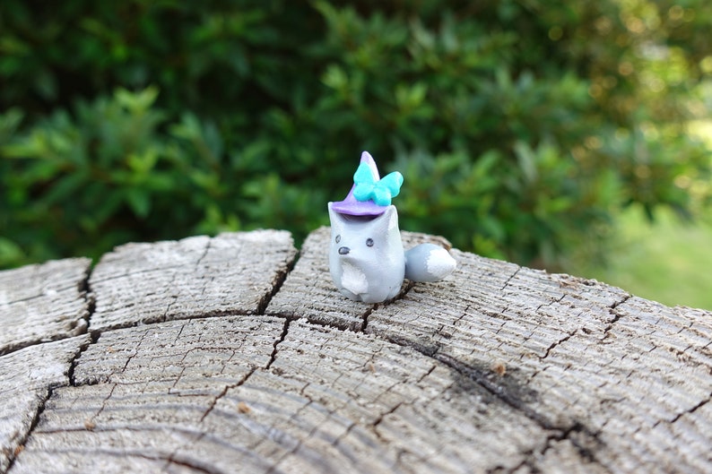 May include: A small gray clay figurine of a fox wearing a purple and blue witch hat with a blue butterfly on top. The figurine is sitting on a gray wooden log.