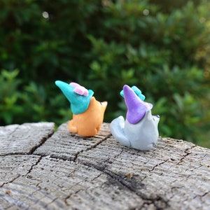 May include: Two small, handmade figurines of animals wearing hats. The figurine on the left is orange and wearing a teal hat with a pink bow. The figurine on the right is gray and wearing a purple hat.