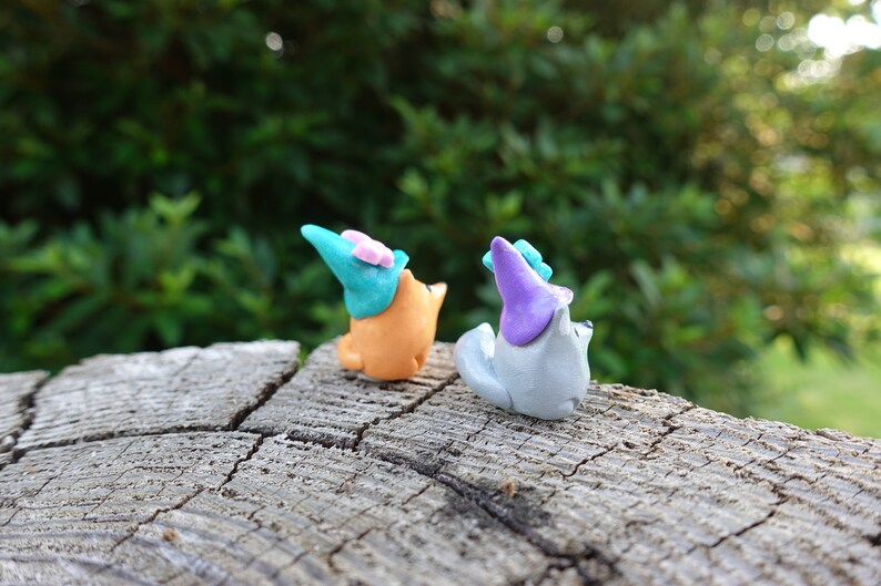 May include: Two small, handmade figurines of animals wearing hats. The figurine on the left is orange and wearing a teal hat with a pink bow. The figurine on the right is gray and wearing a purple hat.