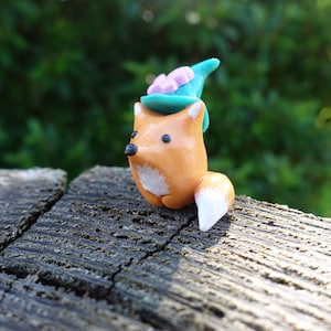 May include: A small orange fox figurine with a white tail and a teal blue hat with pink flowers on top. The fox is sitting on a piece of weathered wood.