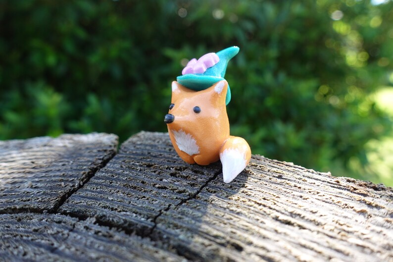 May include: A small orange fox figurine with a white tail and a teal blue hat with pink flowers on top. The fox is sitting on a piece of weathered wood.