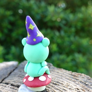 May include: A green frog wearing a purple and yellow star-patterned wizard hat sits on a red and white polka-dotted mushroom.