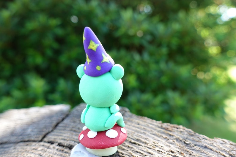 May include: A green frog wearing a purple and yellow star-patterned wizard hat sits on a red and white polka-dotted mushroom.