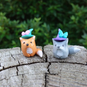 May include: Two small clay figurines of foxes wearing witch hats with butterfly accents. The fox on the left is orange and the fox on the right is gray. They are sitting on a wooden surface.