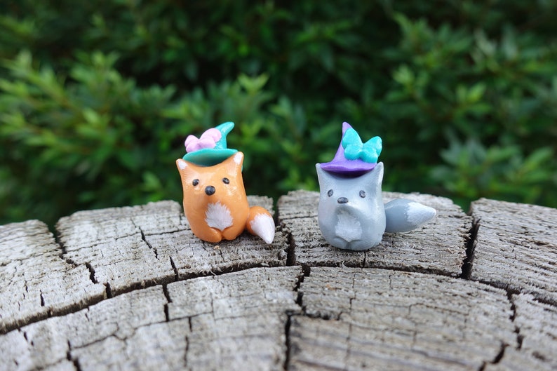 May include: Two small clay figurines of foxes wearing witch hats with butterfly accents. The fox on the left is orange and the fox on the right is gray. They are sitting on a wooden surface.