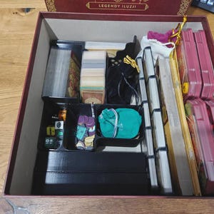 May include: Open board game box with various components. Includes stacks of cards, dice, game pieces, and rule books. The box is burgundy with gold accents and the text "Legendy Iluzji".