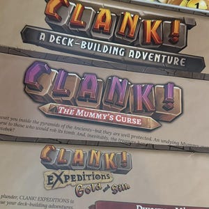 May include: A board game collection featuring Clank! A Deck-Building Adventure, The Mummy's Curse expansion, and Expeditions Gold and Silk. The set includes gold-colored circular tokens with numerical values stored in white plastic trays.