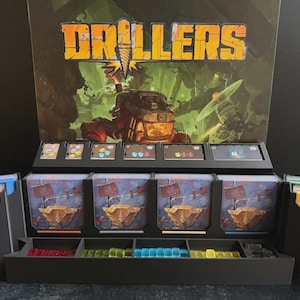 May include: The board game Drillers features a black plastic organizer containing stacks of illustrated cards, small translucent red, green, blue, and yellow cubes, and a large vertical backboard displaying the game title and a mining machine.