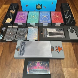 May include: A board game set with various components, including cards, tokens, and a game board. The game box is blue with the text "Old King's Crown." The cards are in purple, green, blue, and red. The set is arranged on a wooden surface.