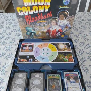 May include: A Moon Colony Bloodbath board game box and open game tray. The box cover features two astronauts and a robot on a lunar surface. The game tray contains a circular player board with sections for research, farm, and build, plus card decks.