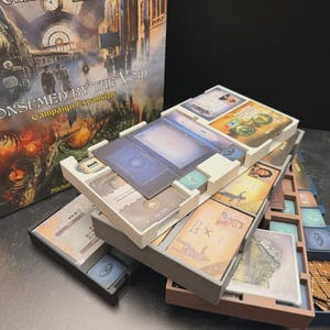 Puede incluir: Una caja de juego de mesa titulada Consumed by the Void Campaign Expansion junto a una bandeja organizadora de plástico blanco y gris de varios niveles, llena de varias cartas de juego rectangulares, fichas cuadradas pequeñas y piezas de madera.