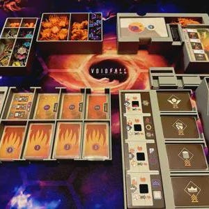 May include: A board game setup with various components. There are several gray trays filled with cards, tokens, and other game pieces. The game board features a central design with the word "VOIDFALL". The cards have flame and skull graphics.