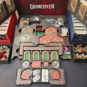 May include: A board game organizer for Grimcoven, featuring various compartments for cards, tokens, and game pieces. The organizer is black and gray, with red and green accents. The game box is open, revealing the game's components.