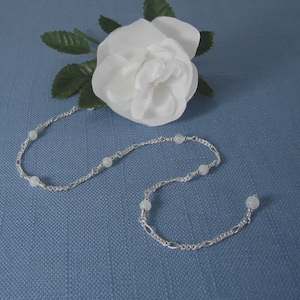 Op de afbeelding: Een zilveren ketting met witte kralen. De ketting ligt op een blauwe achtergrond.