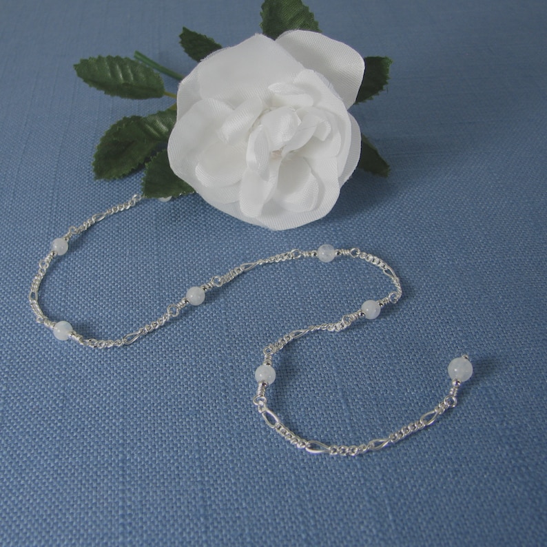 Op de afbeelding: Een zilveren ketting met witte kralen. De ketting ligt op een blauwe achtergrond.