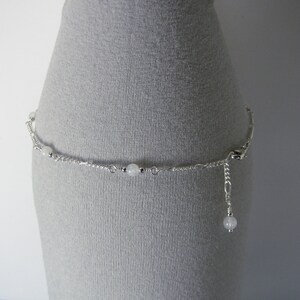 Op de afbeelding: Een zilveren enkelketting met witte kralen. De enkelketting heeft een sluiting en een kleine kraal die aan het einde hangt.