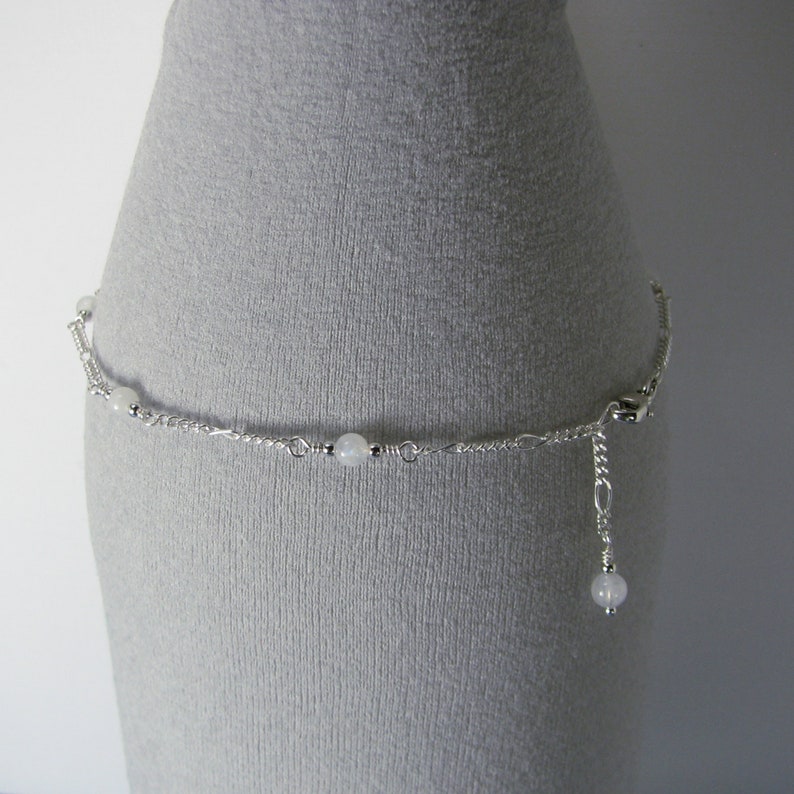 Op de afbeelding: Een zilveren enkelketting met witte kralen. De enkelketting heeft een sluiting en een kleine kraal die aan het einde hangt.