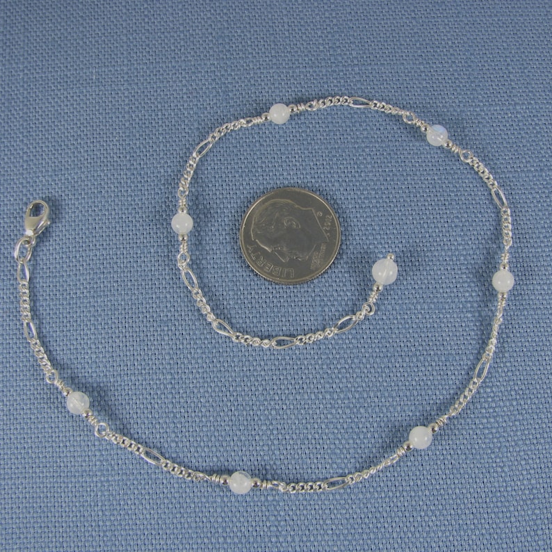 Op de afbeelding: Een zilveren enkelketting met witte maansteen kralen. De enkelketting is ongeveer zo groot als een dubbeltje.