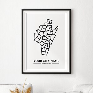 Op de afbeelding: Zwart-witte lijntekening van een stadskaart met de tekst "YOUR CITY NAME" en "YOUR COUNTRY" onder de kaart. De print is ingelijst in een zwart frame en hangt aan een witte muur boven een witte bank met kussens.
