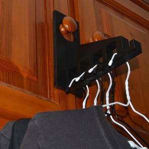 May include: A black wooden wall-mounted clothes hanger with a series of slots for hangers. The hanger has three wooden knobs and is mounted on a wooden door. White hangers are in the slots, holding black shirts.