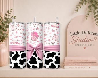 May include: Three white tumblers with a pink cowgirl theme. The design includes cowboy boots, hats, stars, and hearts. The bottom section has a black and white cow print, and a pink bow is in the middle. The tumblers have a straw.