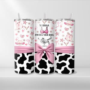 May include: Three stainless steel tumblers with a cow print design. The tumblers have a pink ribbon and a pink design with the text "Yeehaa Swing Yer Bra".